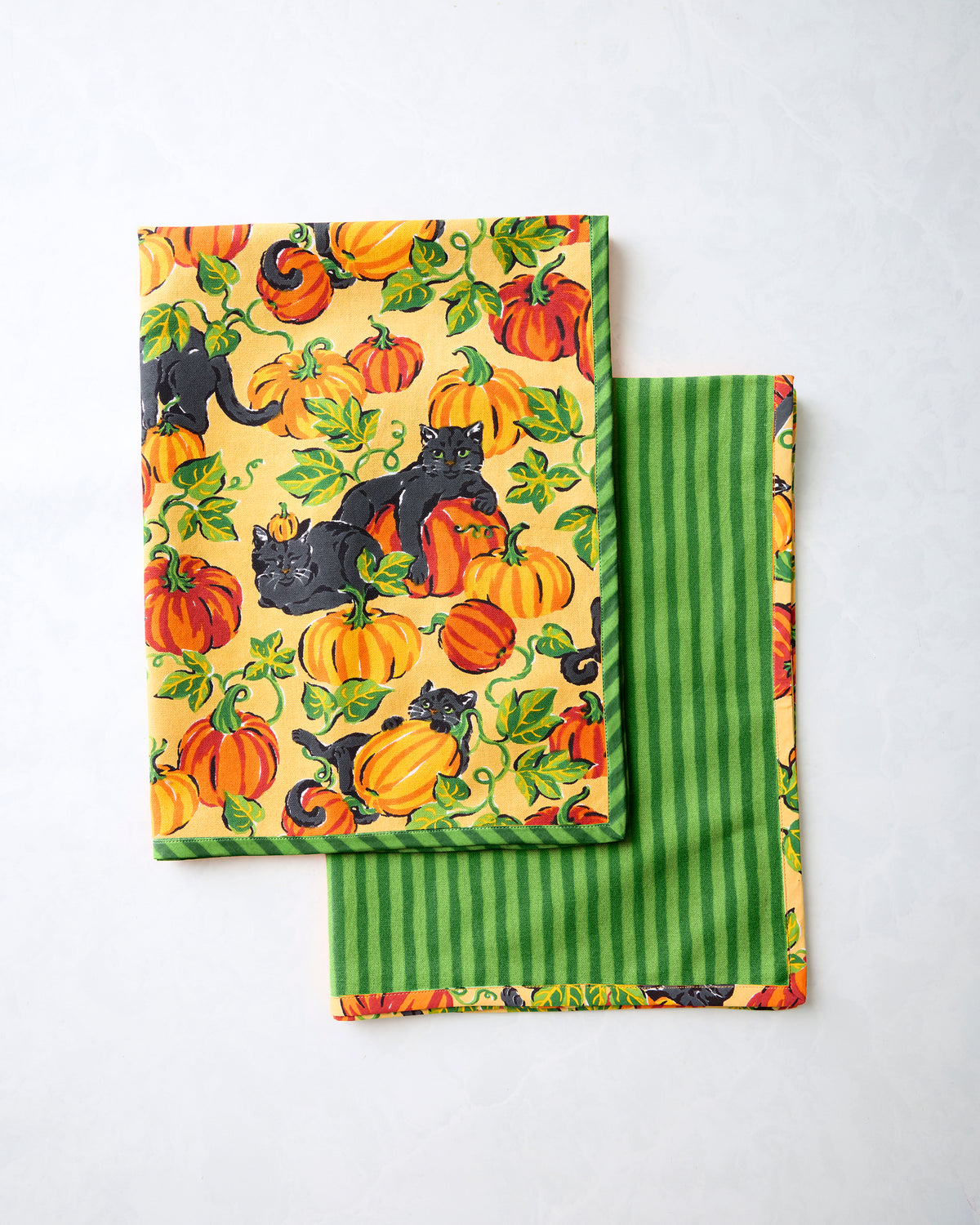 Two folded tea towels showing the pumpkin-and-black-cat print and a green striped reverse side.