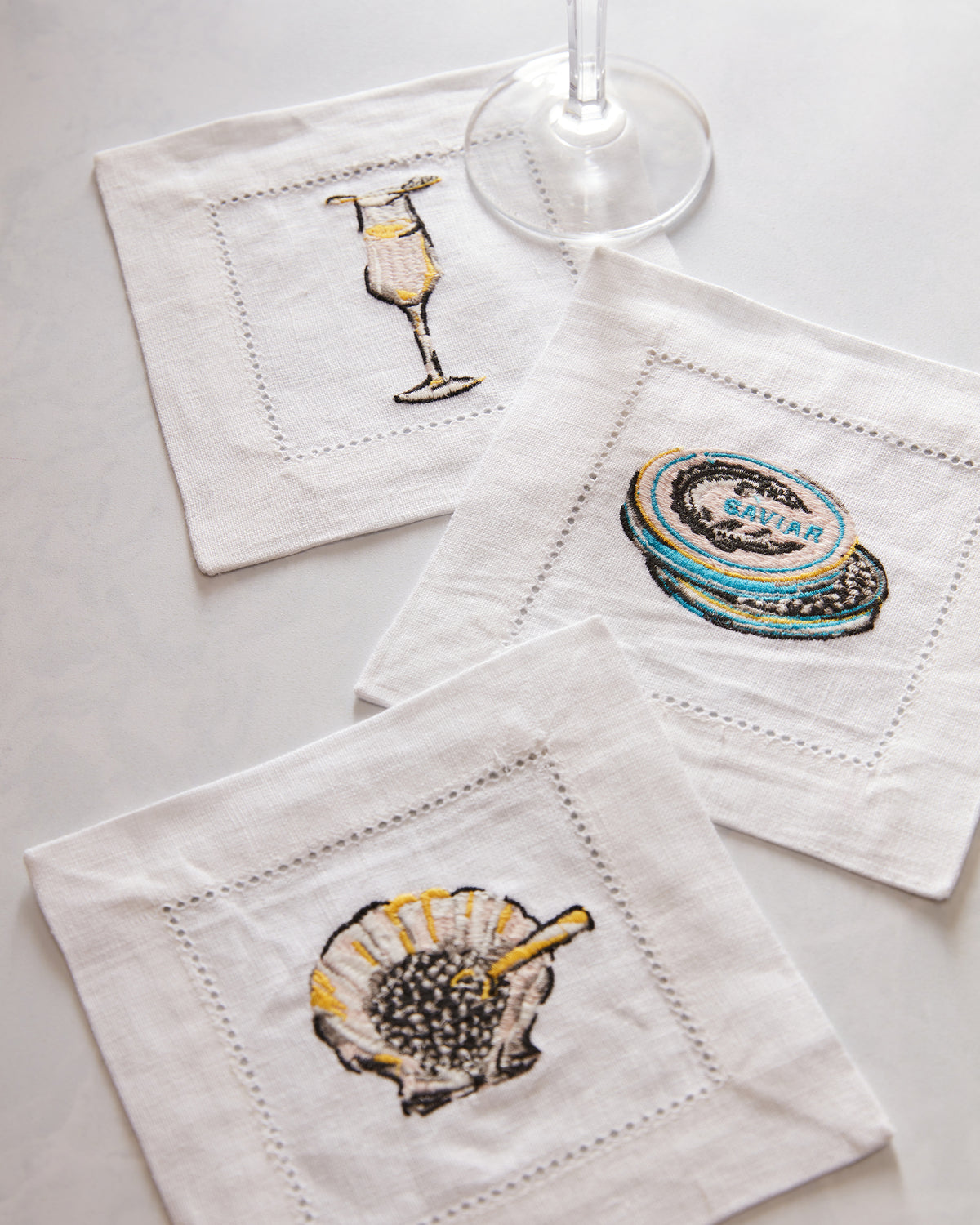 Three white Champagne & Caviar cocktail napkins arranged on a surface, embroidered with a champagne glass, caviar tin, and caviar; a wine glass is nearby.