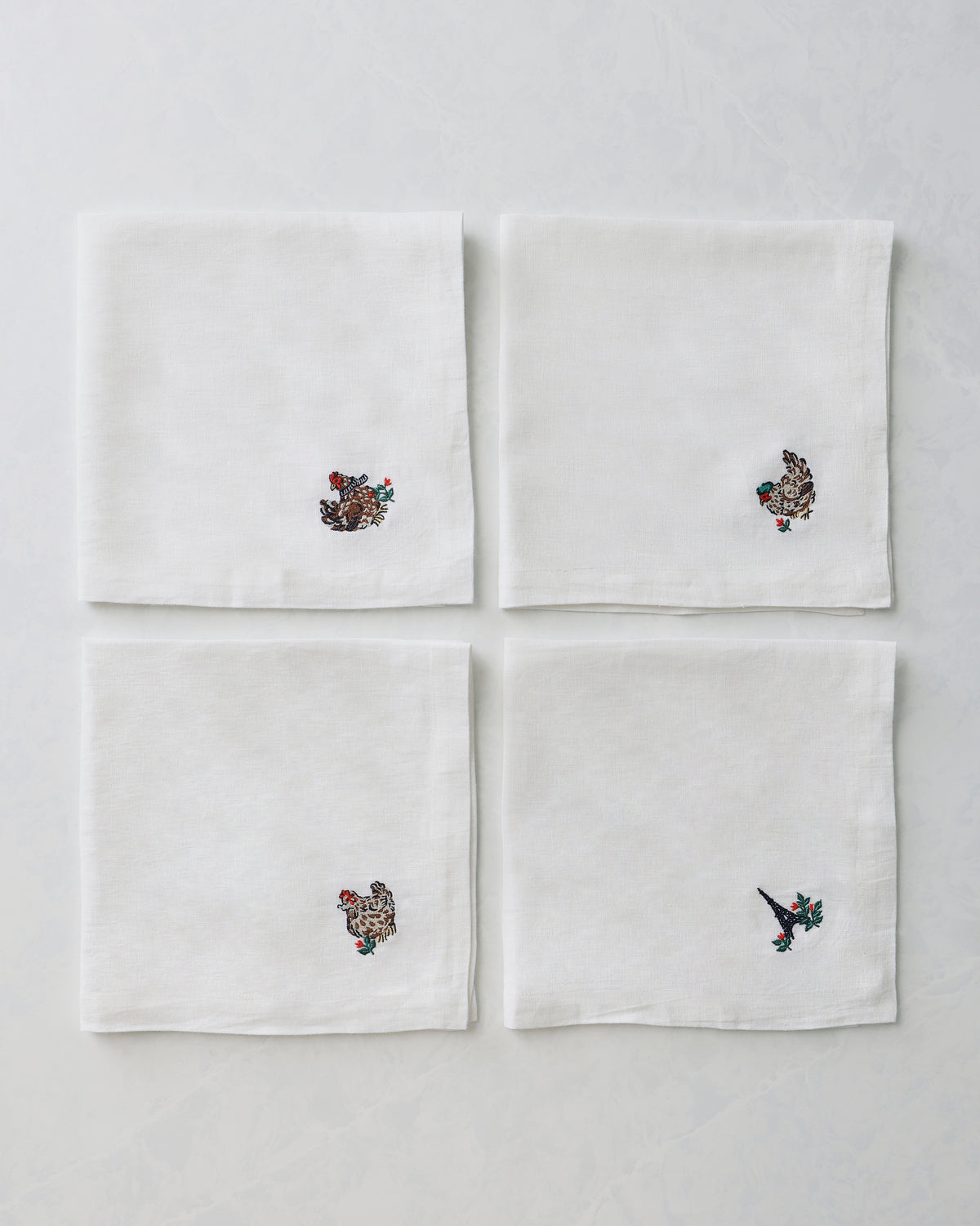 Four white dinner napkins arranged in a square, each with a small embroidered holiday motif near one corner.