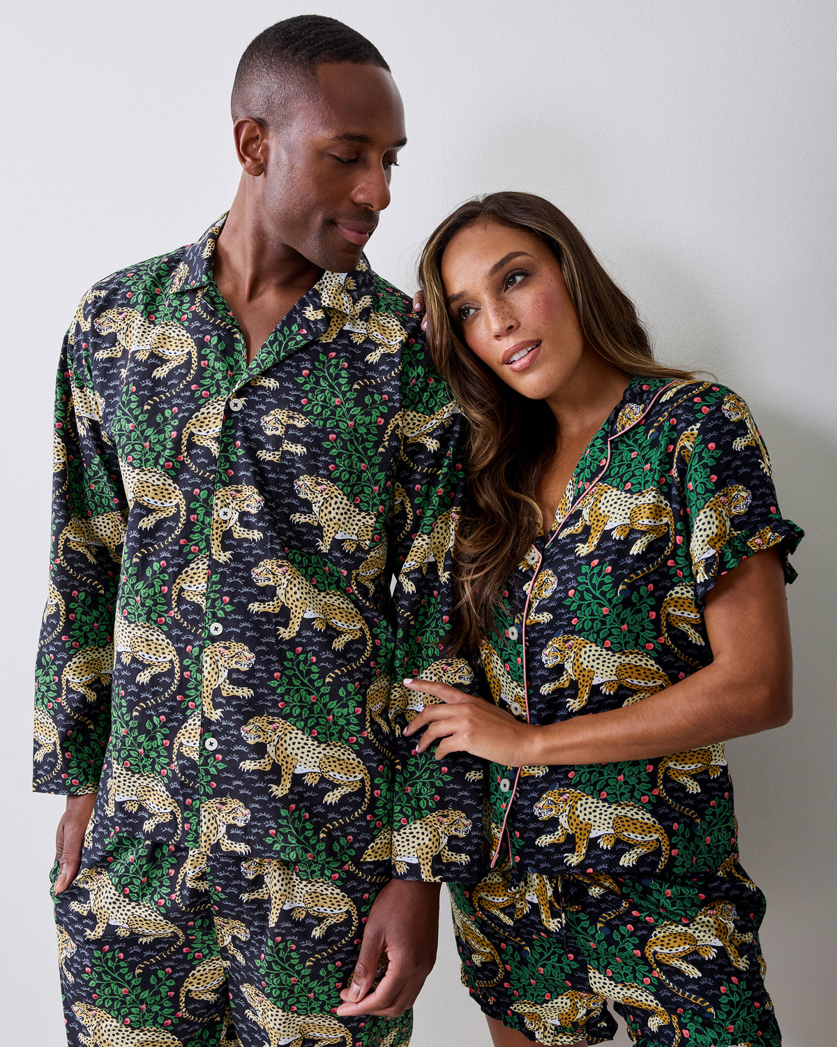 A person wears a long-sleeve button-front pajama set with a jungle-cat and leafy-vine print; another person wears a coordinating pajama top and shorts.
