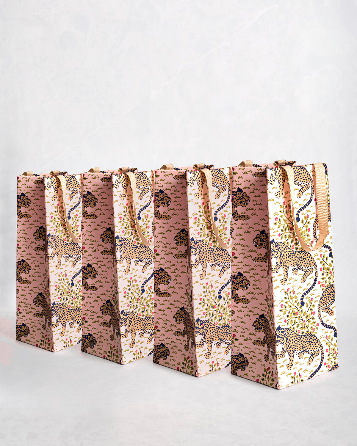 Set of four paper wine gift bags with blush, cream, and leopard-patterned designs and sparkly handles.