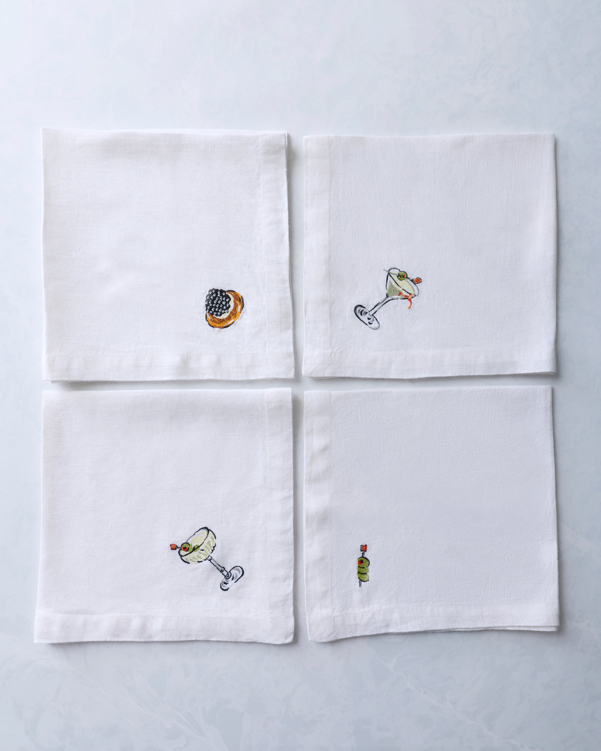 Four white dinner napkins arranged in a grid, each with a small embroidered food or cocktail motif.