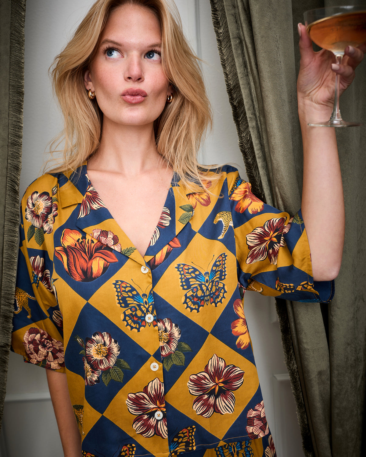 Close view of a person wearing the Golden Hour Botanical Checkerboard short-sleeved button-front top with floral and butterfly motifs, holding a glass.