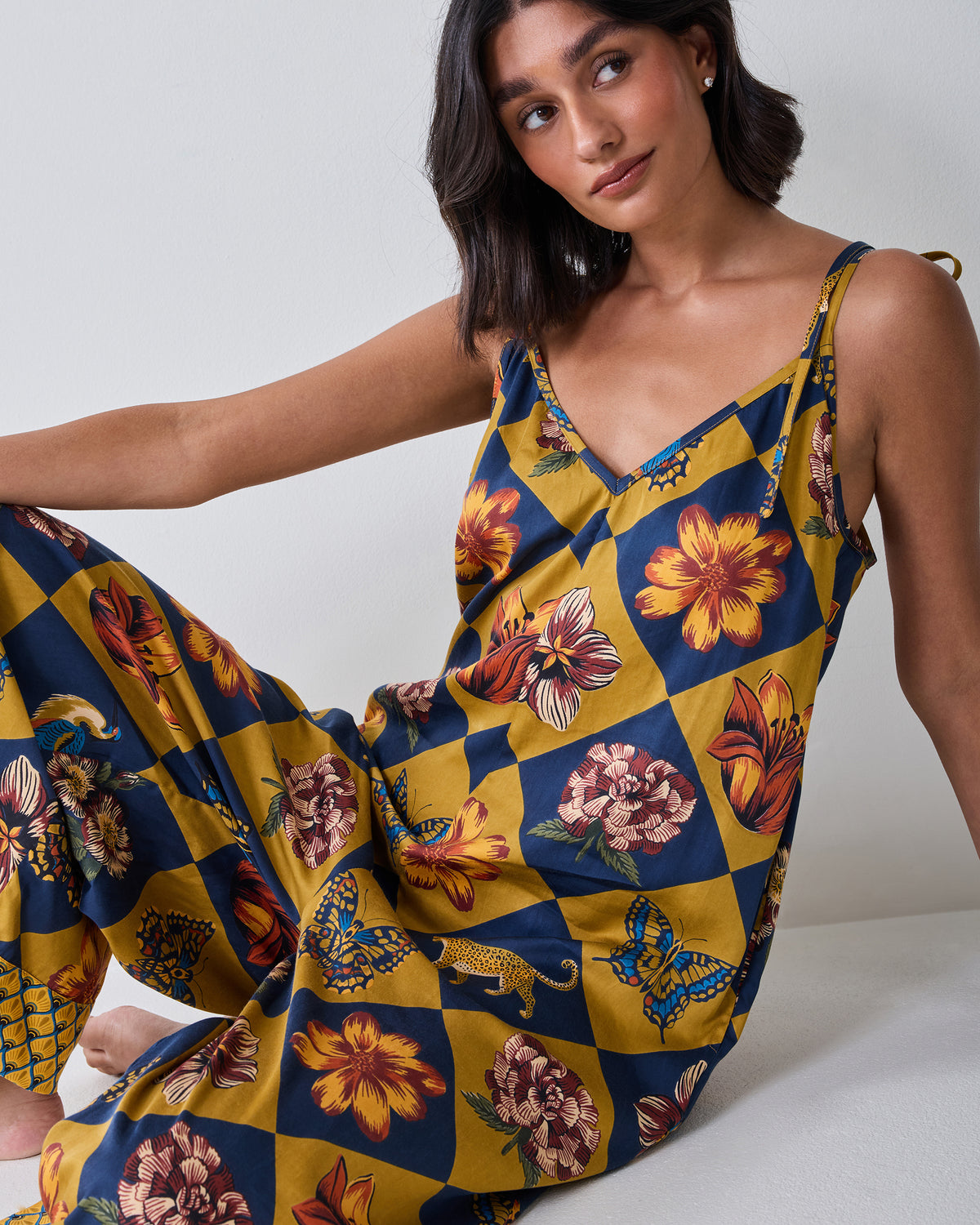 Seated model wearing the Botanical Checkerboard Jaipur jumpsuit, showing the V-neckline, tie shoulder straps, wide legs, and patterned print.