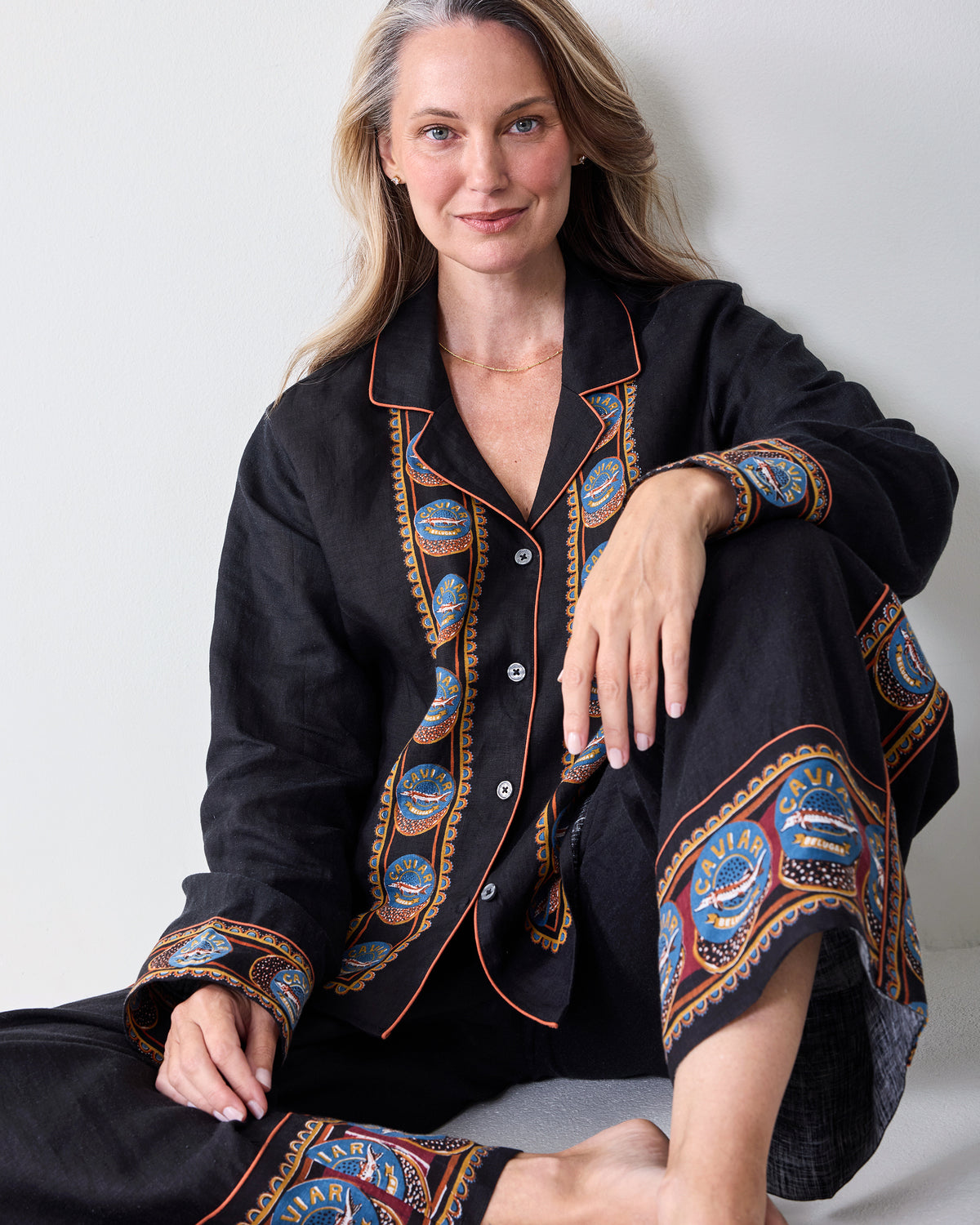 Seated view of the black tunic and wide-leg pants with decorative patterned trim on the front, cuffs, and pant legs.