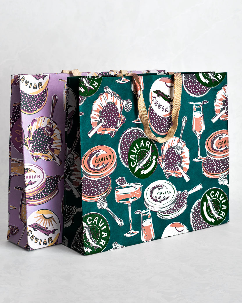 Two paper gift bags with Champagne & Caviar print and sparkly handles, one lavender and one teal.