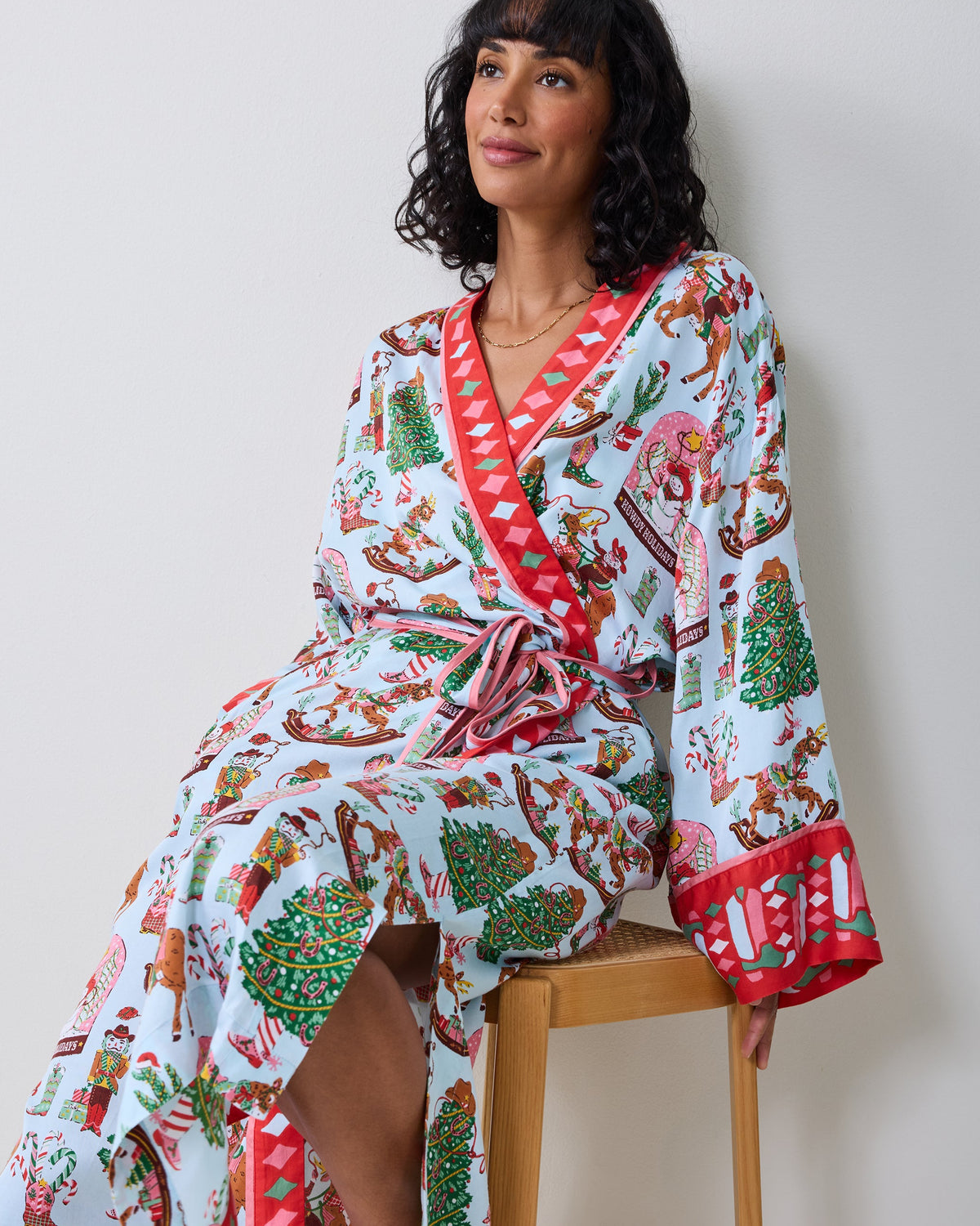 Person wearing a light-colored long wrap robe with a contrasting red trim and a Cowboy Christmas print of Santa, trees, boots, and candy canes.