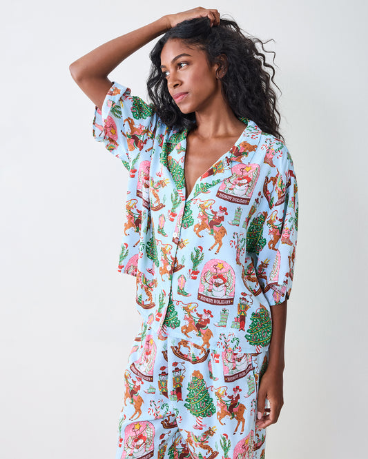 Model wearing the pale blue Cowboy Christmas pajama set with a button-front top and wide-leg pants, printed with Santa riding reindeer, Christmas trees, boots, and candy canes.