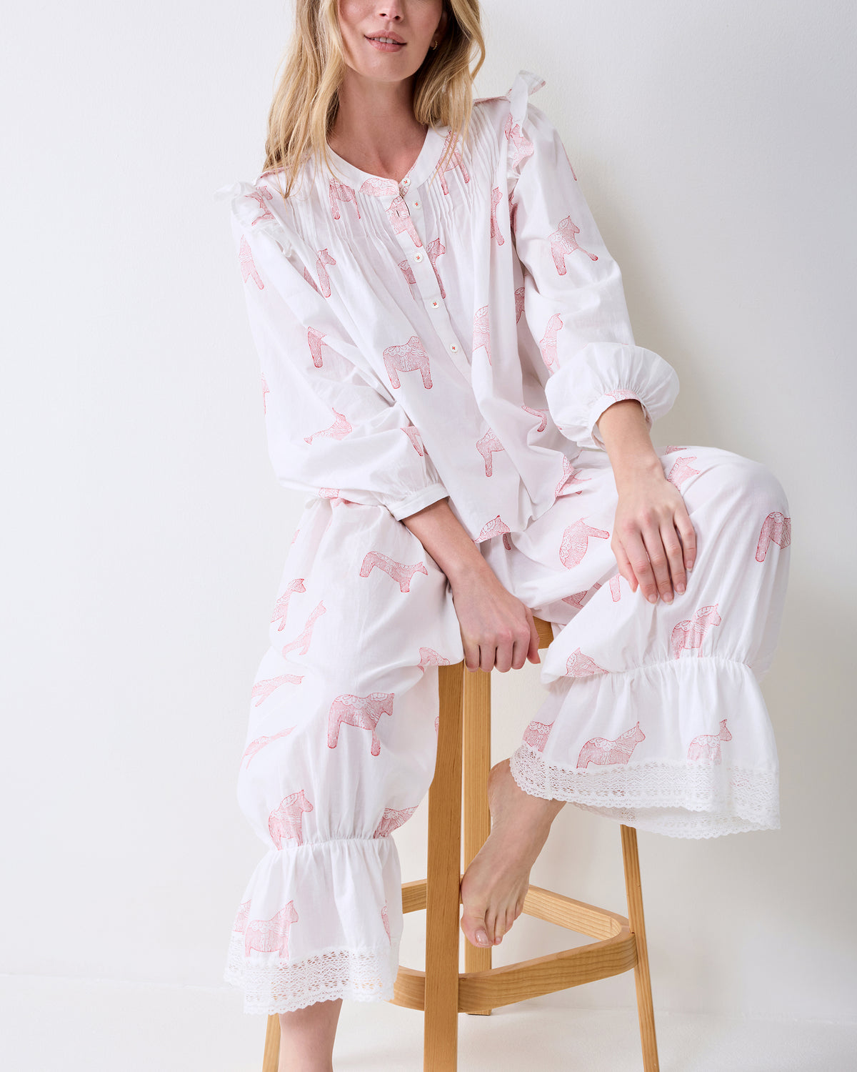 Person seated on a wooden stool wearing a white pajama set with pink horse prints and lace-trimmed hems.
