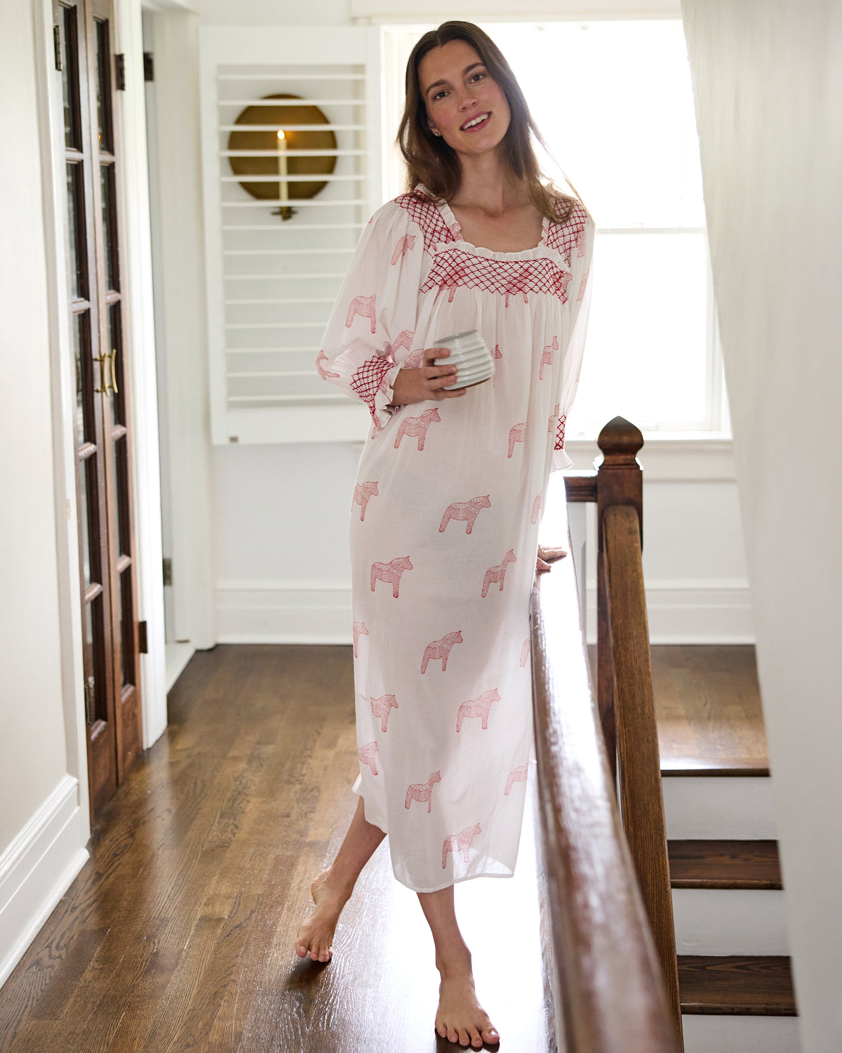 Front view of a long white nightgown with pink horse prints and red smocked trim, worn on a person standing by a staircase.