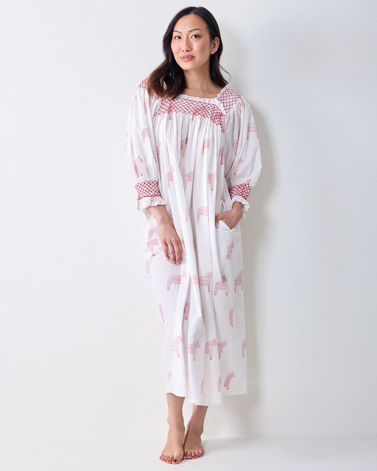 Front view of a person wearing a long white nightgown with pink horse prints, red smocked trim, and side pockets.