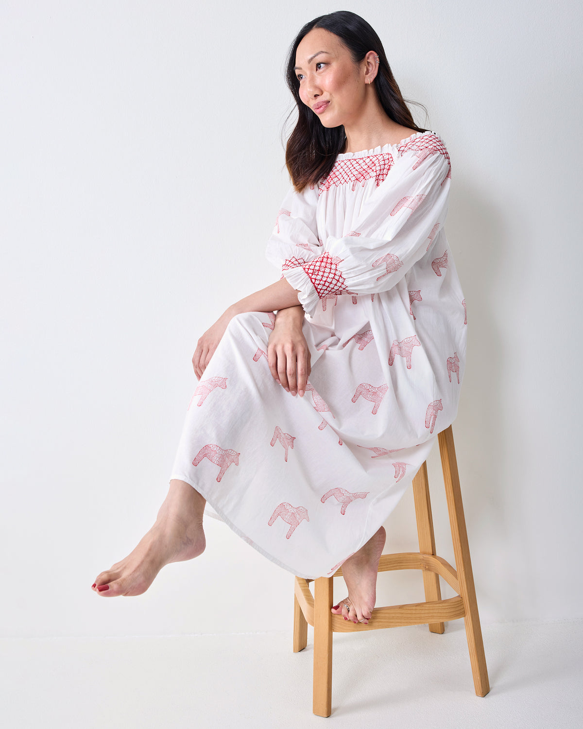 Side view of a person seated on a wooden chair wearing a long white nightgown with pink horse prints and red smocked trim.