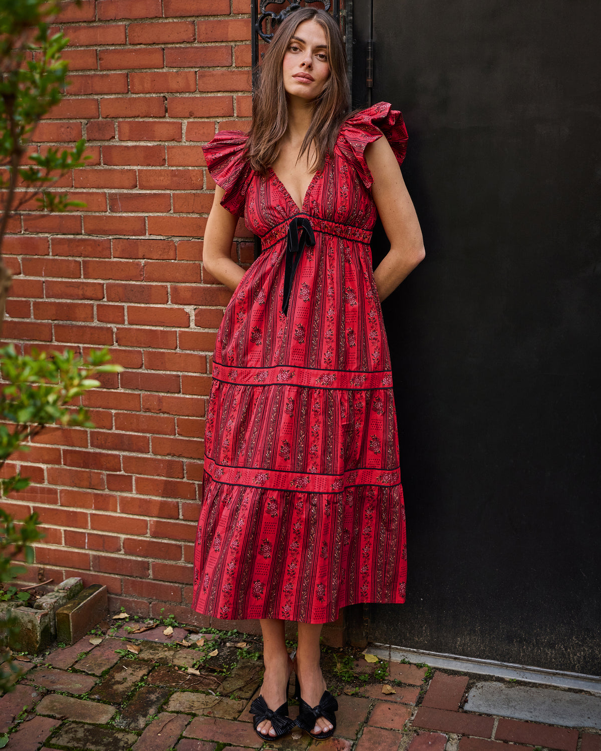 Front view of the Festive Roses Cecily Dress in Heirloom Red, showing its ruffled sleeves, waist tie, V-neckline, and tiered skirt outdoors.