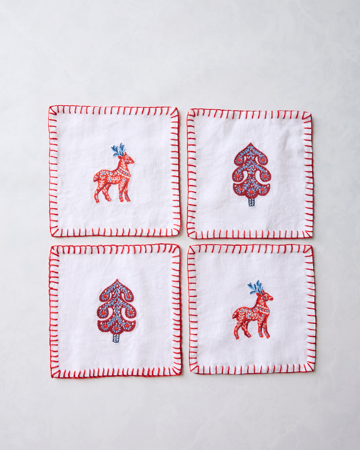 Four white square textiles arranged in a grid, each with a red stitched border and a central red-and-blue deer or tree motif.