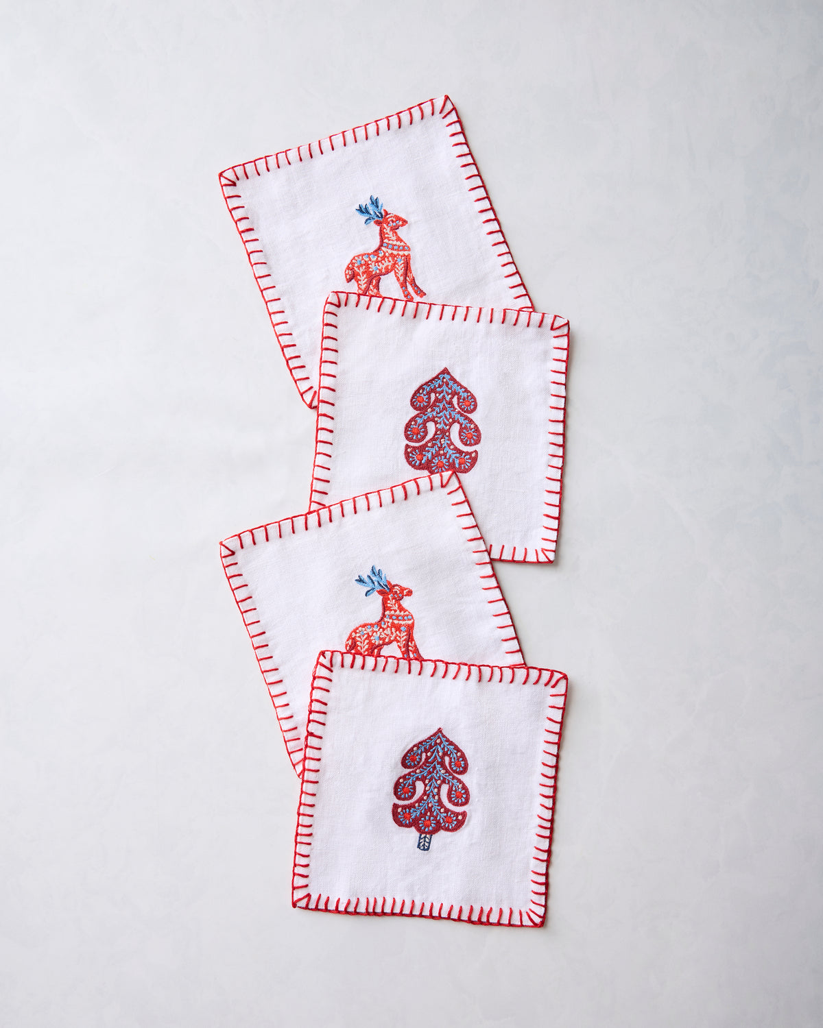 Four white cocktail napkins with red stitched edges, featuring alternating embroidered deer and stylized tree motifs.