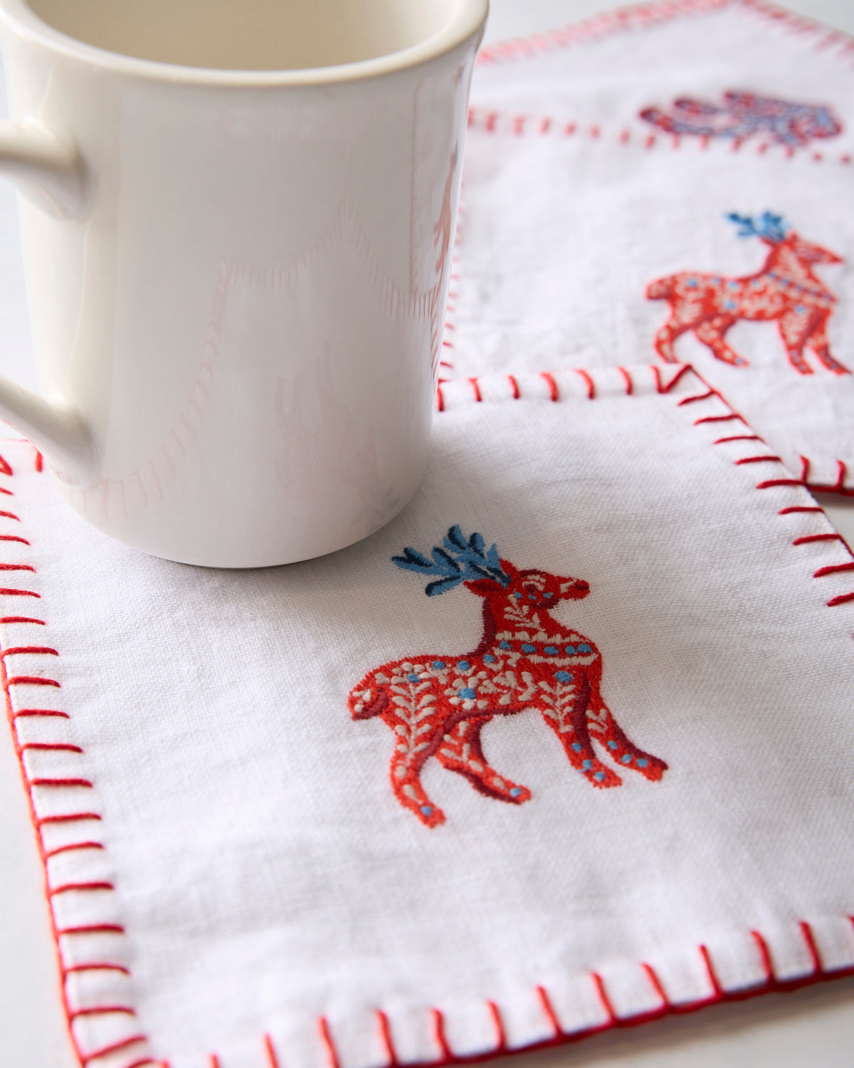White cocktail napkin with an embroidered red deer beside a white mug, with additional embroidered napkins in the background.