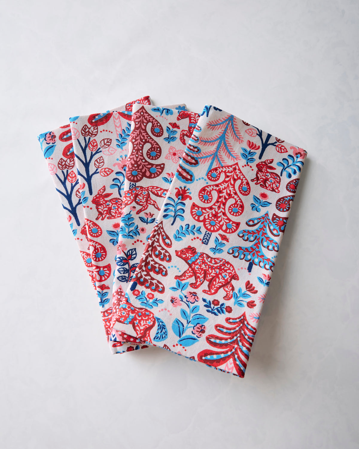 Several folded dinner napkins displayed together, showing the Folklore Forest print of red and blue trees, animals, flowers, and decorative motifs.