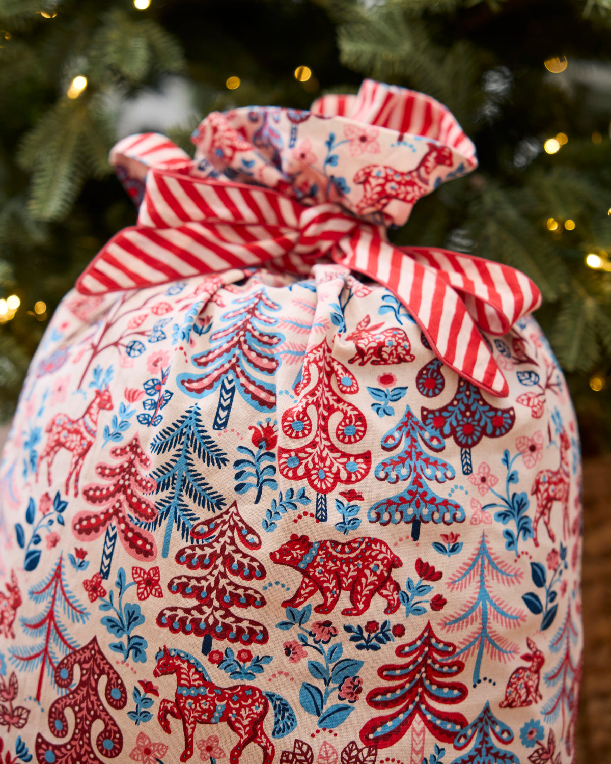 Cream Folklore Forest gift sack with a red-and-blue woodland print, gathered and tied with a red-and-white striped ribbon.