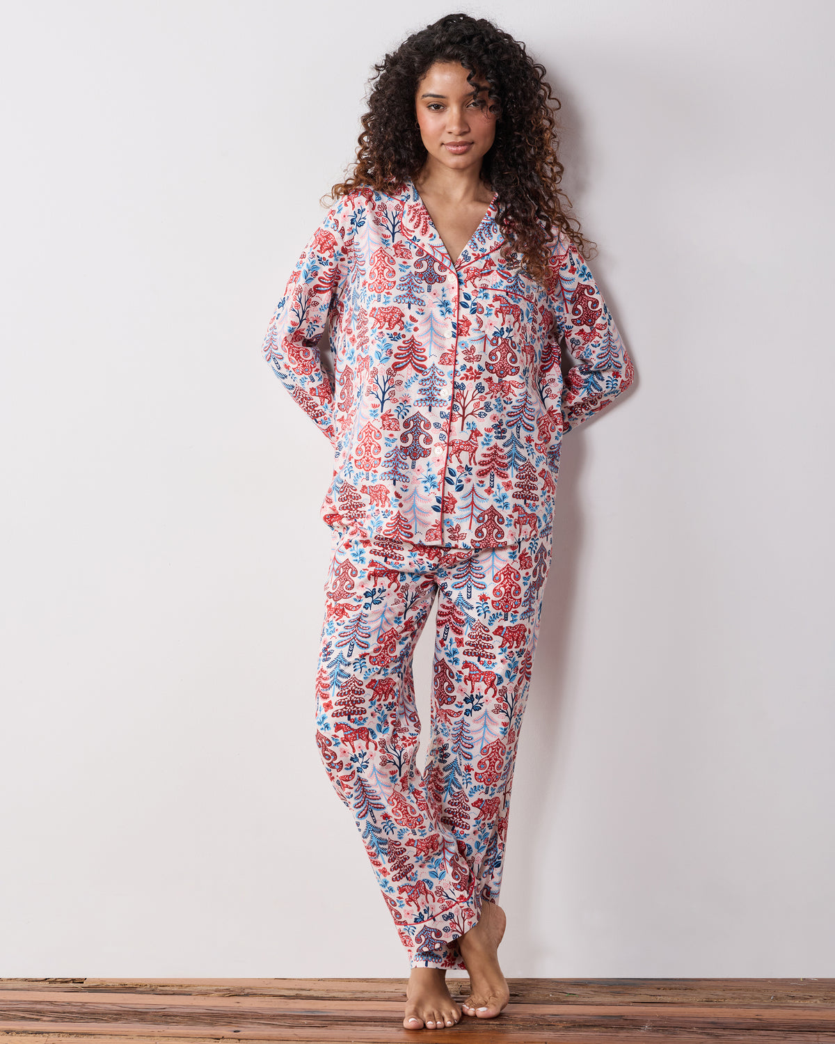 Person wearing a cream long-sleeve button-front pajama top and matching pants with a red-and-blue forest print.