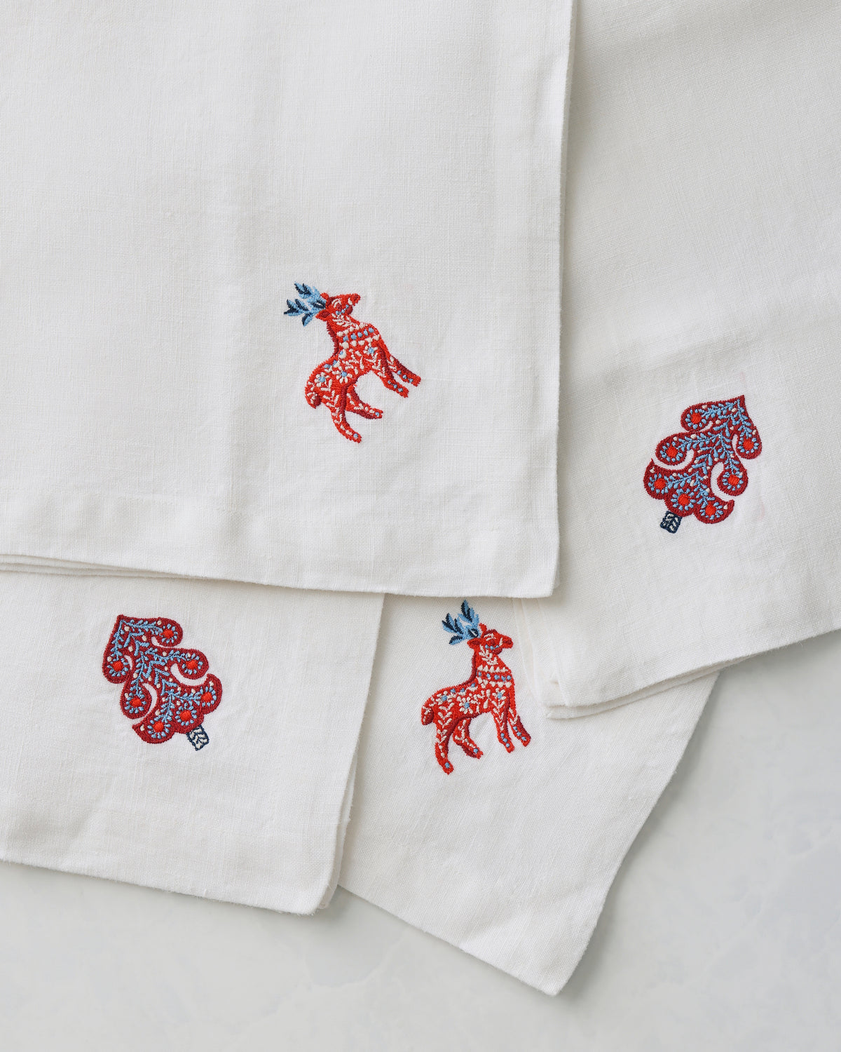 Close-up of four white oversized dinner napkins with embroidered red deer and stylized tree motifs near their corners.