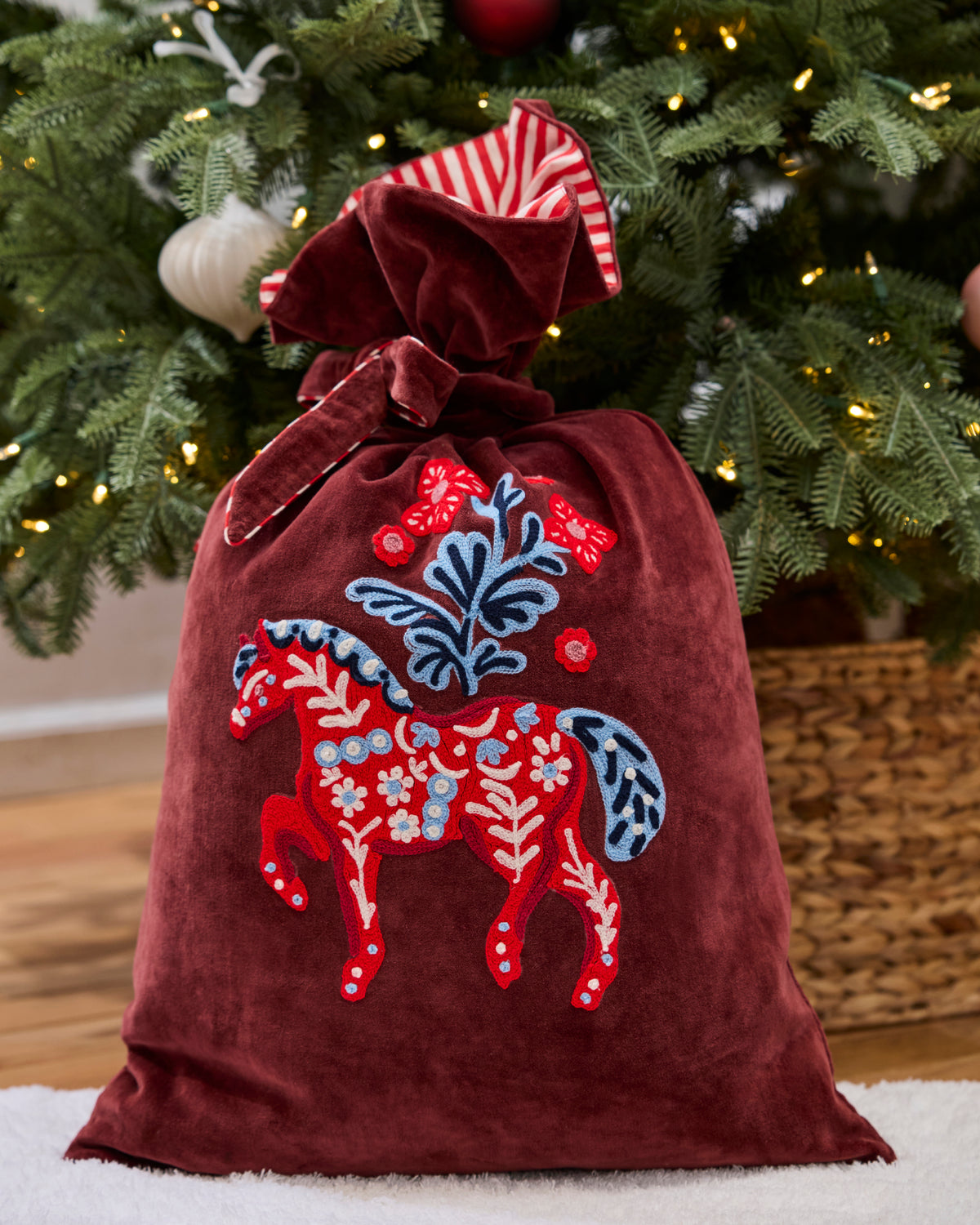 Upright burgundy gift sack with a red embroidered horse and floral motif, shown beside a decorated Christmas tree.