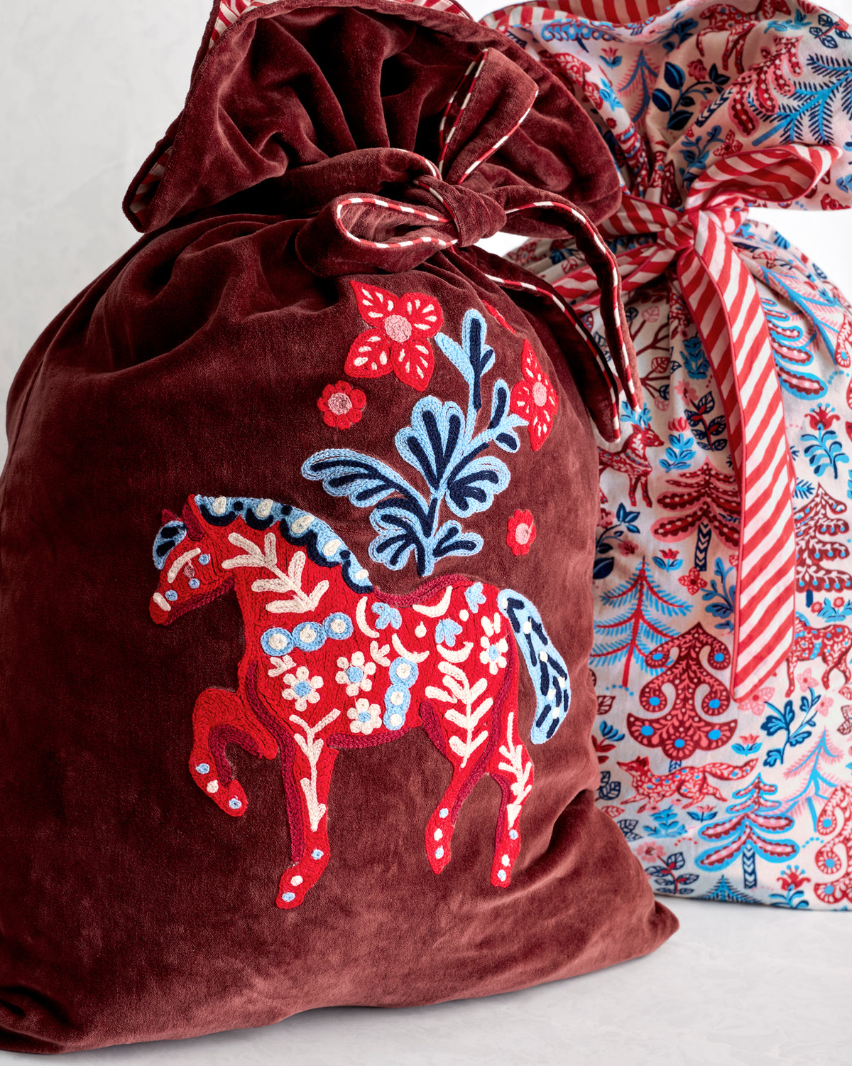 Close-up of a burgundy gift sack with an embroidered horse and floral design, with a patterned sack behind it.