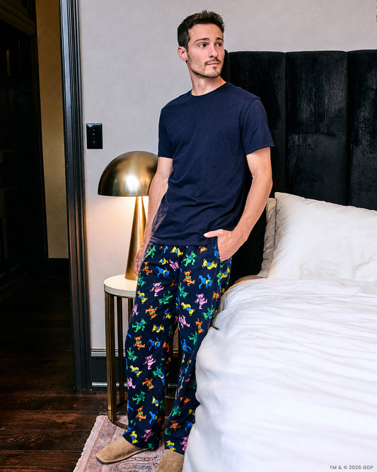 Person wearing the Dancing Bears blue pajama pants with colorful bear figures, standing beside a bed.