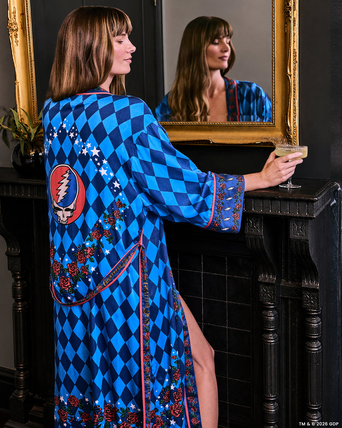 Back view of the blue argyle-print robe with rose and star trim and a large red oval skull-and-lightning graphic, shown in a mirror while holding a drink.