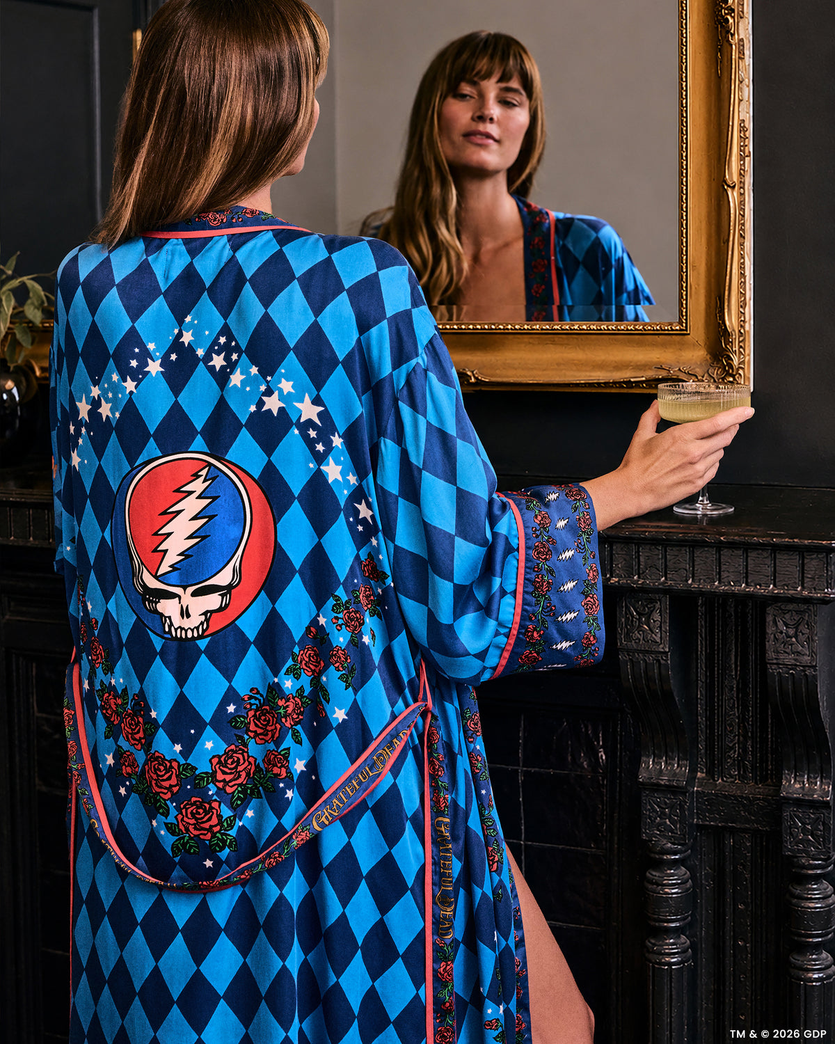 Person wearing a blue diamond-patterned robe with floral trim and a large skull-and-lightning emblem on the back, holding a drink beside a mirror.
