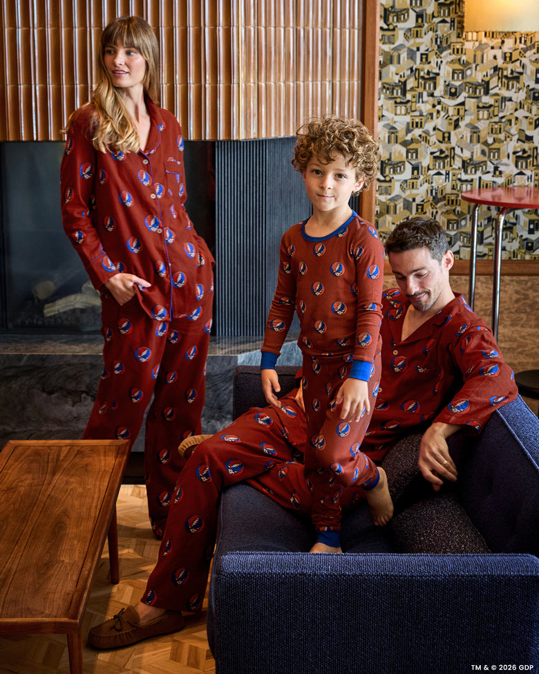 Three people wearing coordinated red patterned pajamas in a living room; one stands while another sits and a child stands in front.