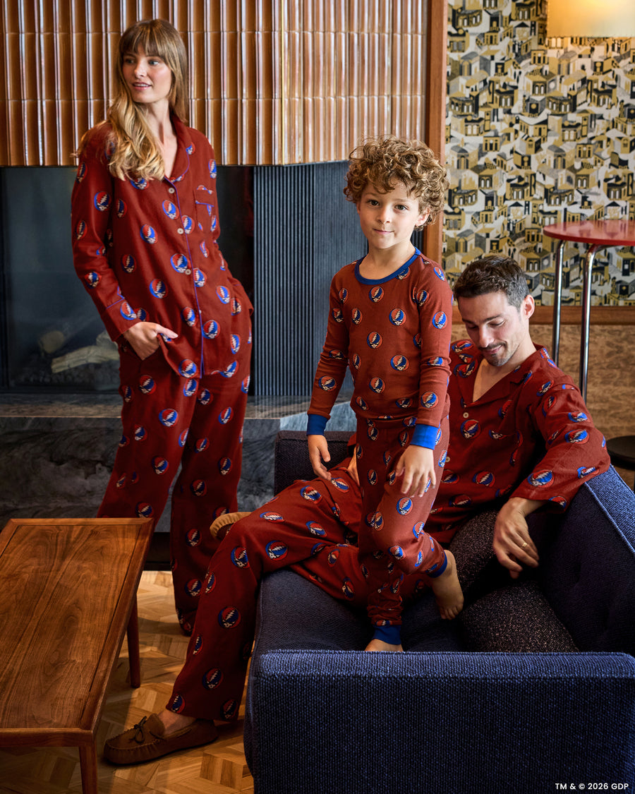 Three people wearing coordinated red patterned pajamas in a living room; one stands while another sits and a child stands in front.