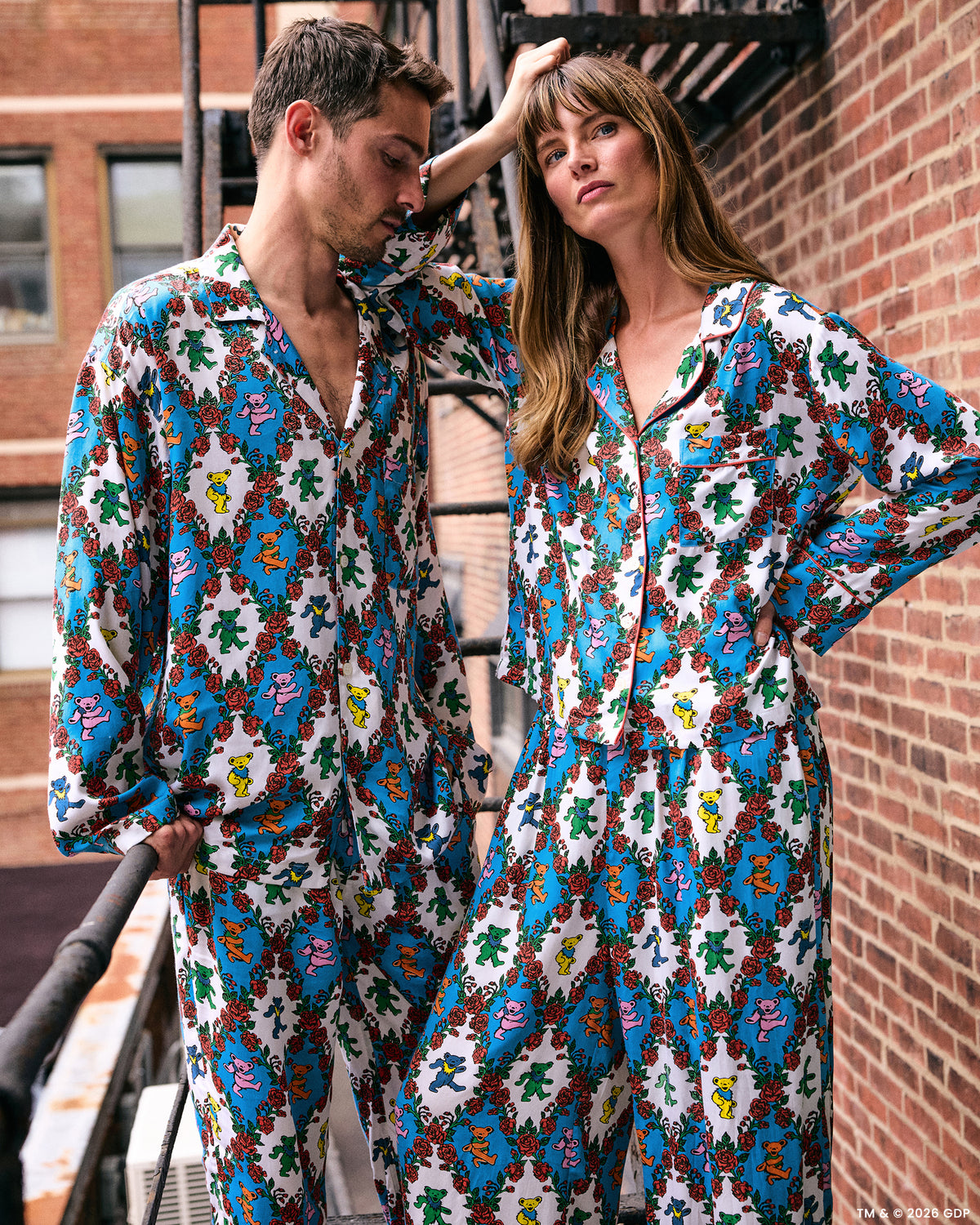 Two people wearing coordinated white pajama sets with colorful figures and red roses beside a brick wall.
