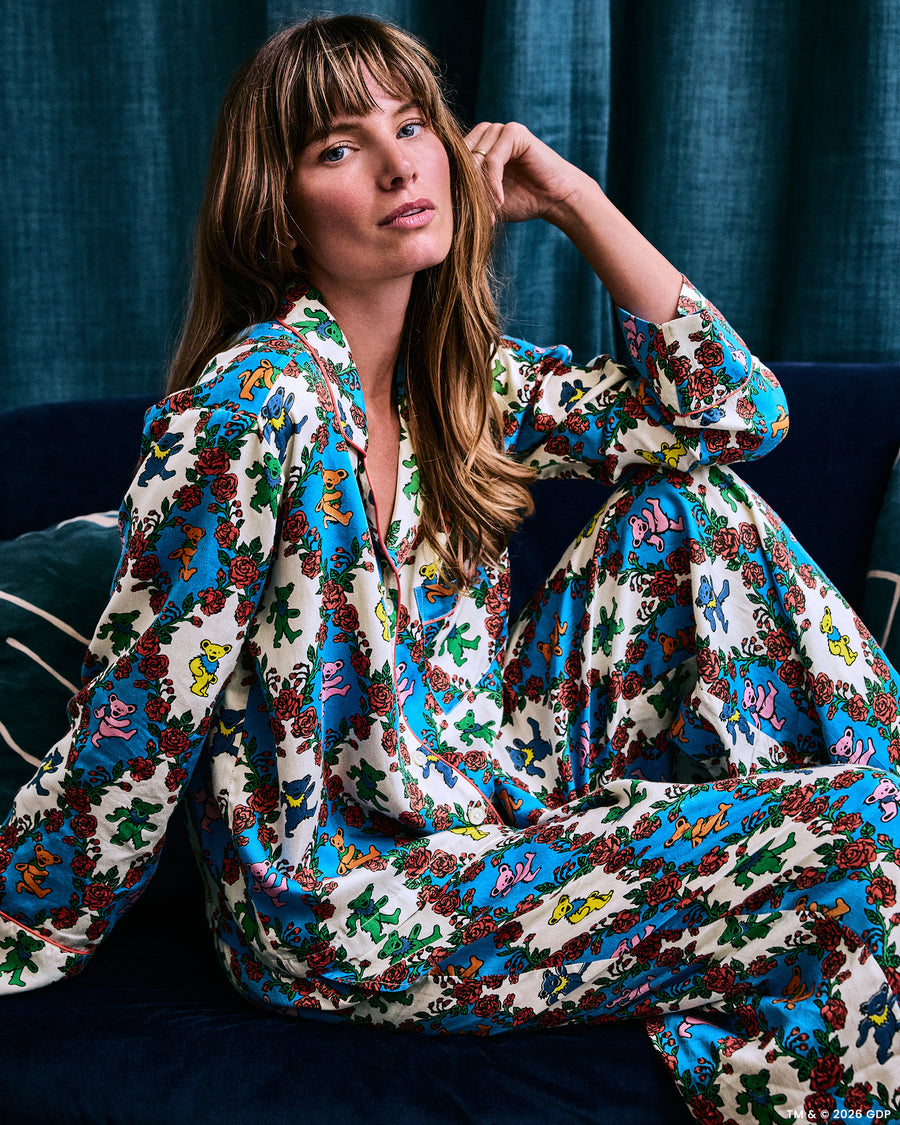 Model seated on a dark sofa wearing a long-sleeved pajama set with a colorful bear-and-rose print.