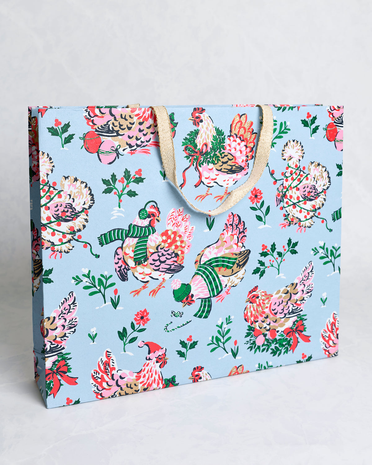 Angled view of the Holiday Hens pale blue handmade gift bag showing its front and side panels with decorated hens, holly branches, berries, and tan handles.