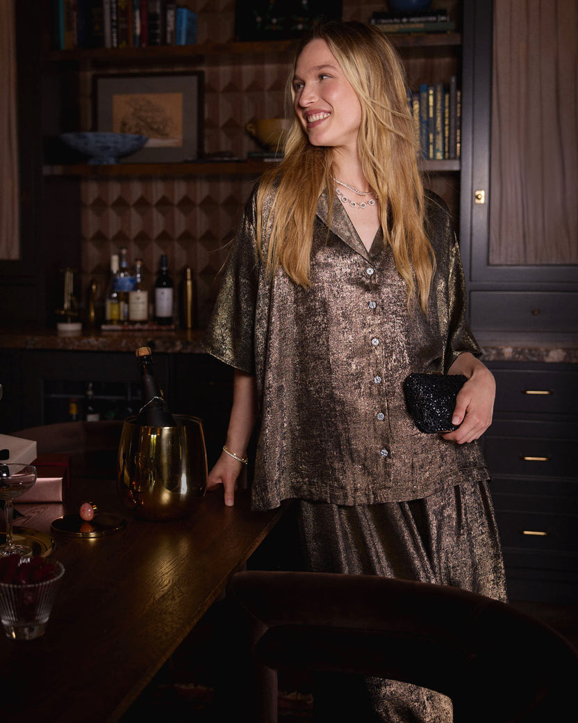 Person wearing a gold-colored short-sleeve pajama shirt and matching skirt, holding a black clutch at a dining table.