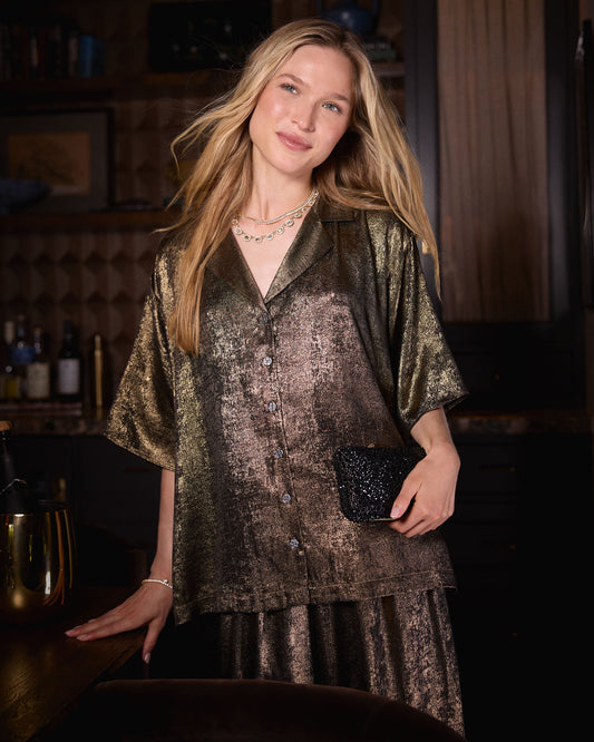 Person wearing the gold Glitzy Lamé Blakely top and matching skirt, holding a black clutch indoors.