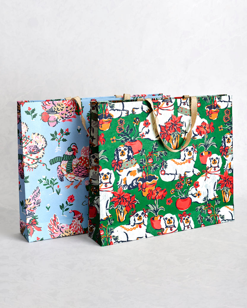 Two paper gift bags, one pale blue with holiday hens and one green with illustrated dogs, flowers, and holiday motifs.