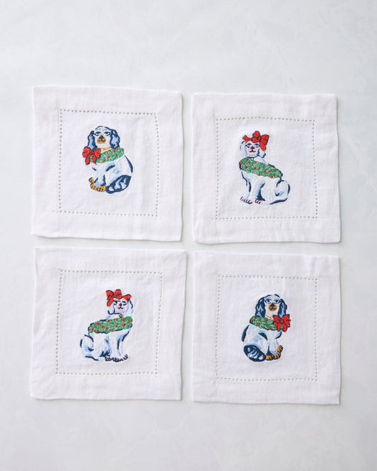Four white cocktail napkins arranged in a square, each embroidered with a Christmas dog wearing a green wreath and red bow.