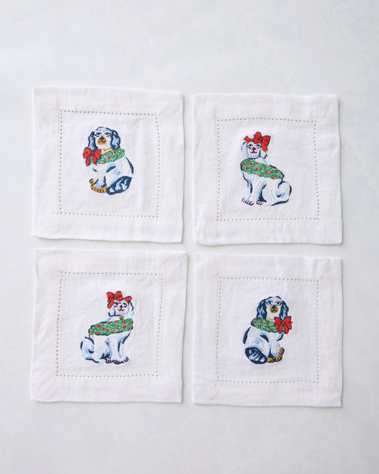 Four white cocktail napkins arranged in a square, each embroidered with a Christmas dog wearing a green wreath and red bow.