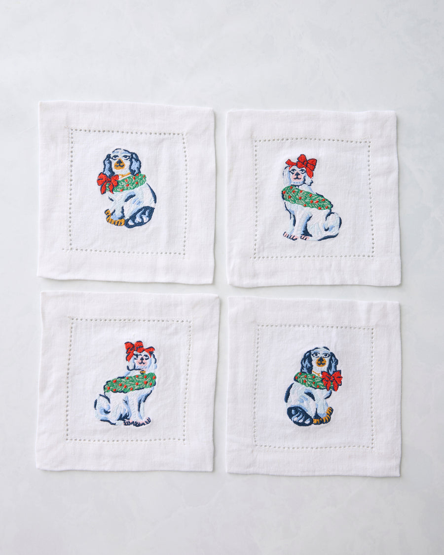 Four white cocktail napkins arranged in a square, each embroidered with a Christmas dog wearing a green wreath and red bow.
