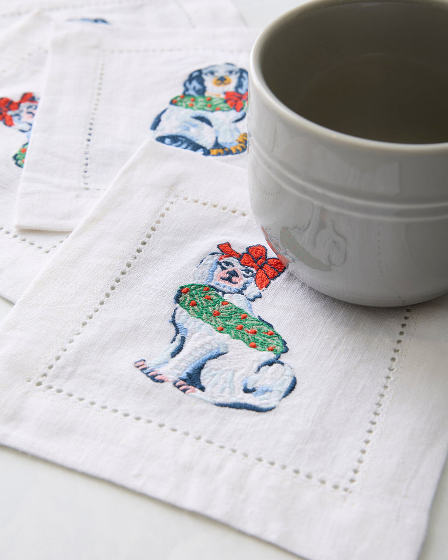 White cocktail napkin embroidered with a Christmas dog, shown beneath a white cup with other napkins in the background.