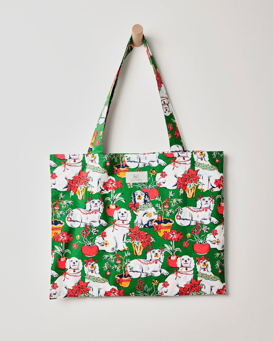 Evergreen Good Dog Christmas tote bag hanging from its handles, with a holiday pattern of white dogs, flowers, plants, and ornaments.