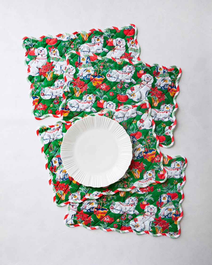 Four scalloped green Christmas placemats with dog, poinsettia, and holiday plant motifs, arranged with a white plate.