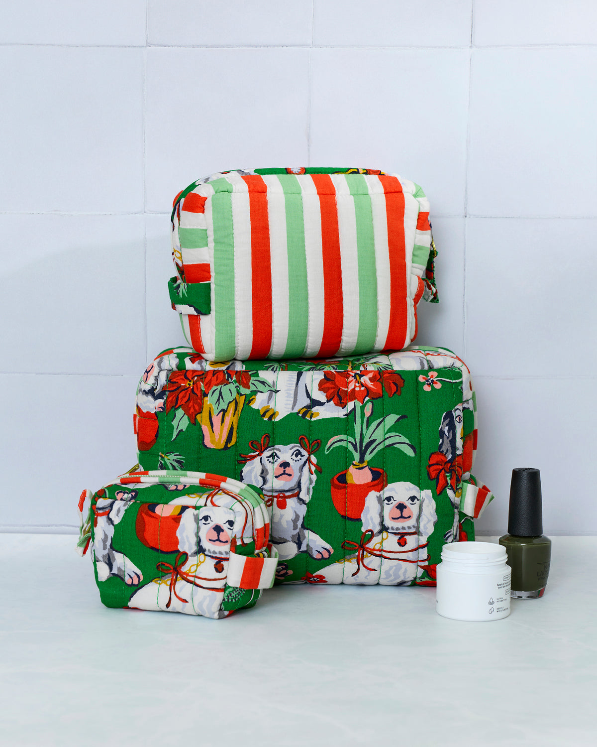 Three quilted evergreen Good Dog Christmas pouches stacked by size, with white dogs, potted plants, and holiday flowers; a striped pouch is on top.
