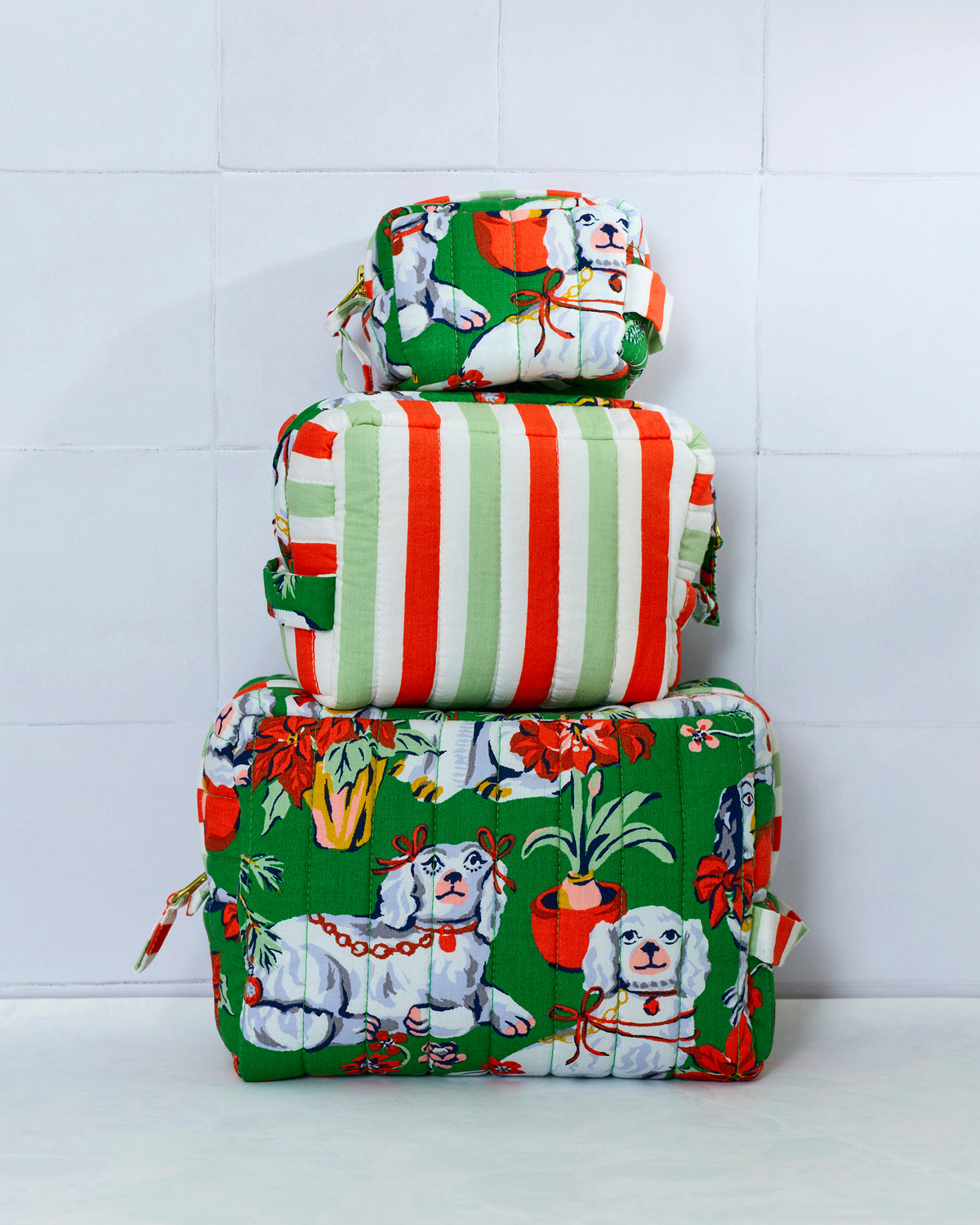 Three quilted evergreen Good Dog Christmas pouches stacked by size, showing white dogs, potted plants, holiday flowers, and the striped back of the middle pouch.