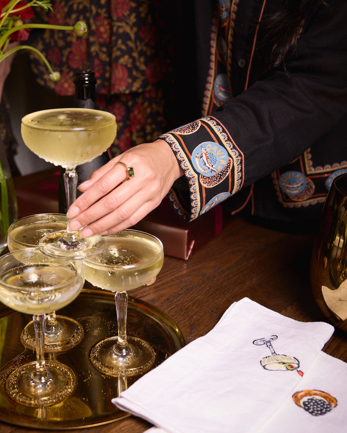 Close-up of a hand beside champagne glasses, showing the circular trim on a black pajama sleeve and embroidered napkins on a tray.