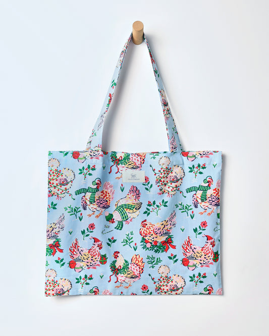 Pale blue Holiday Hens tote bag hanging from its handles, covered with illustrated hens wearing winter hats and scarves.
