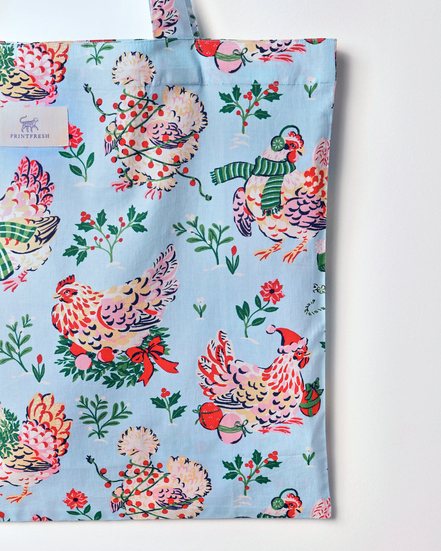 Close view of the Holiday Hens tote’s pale blue print, showing illustrated hens, holly branches, flowers, and holiday accessories.