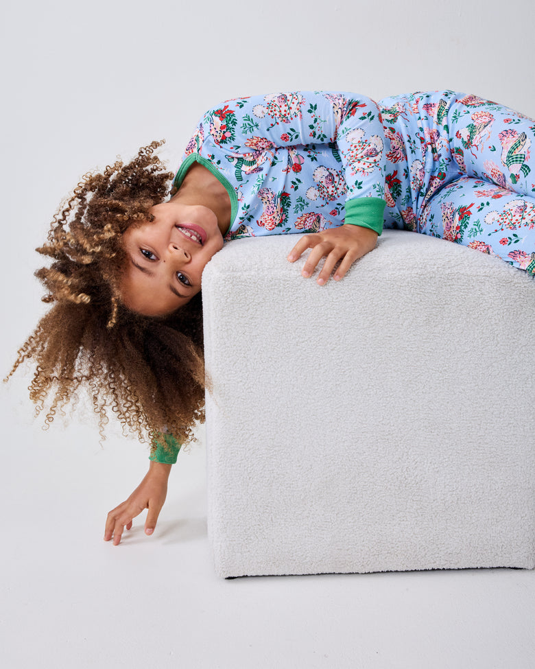 Child in a pale blue Holiday Hens pajama set lies across a white ottoman with their head hanging over the edge.