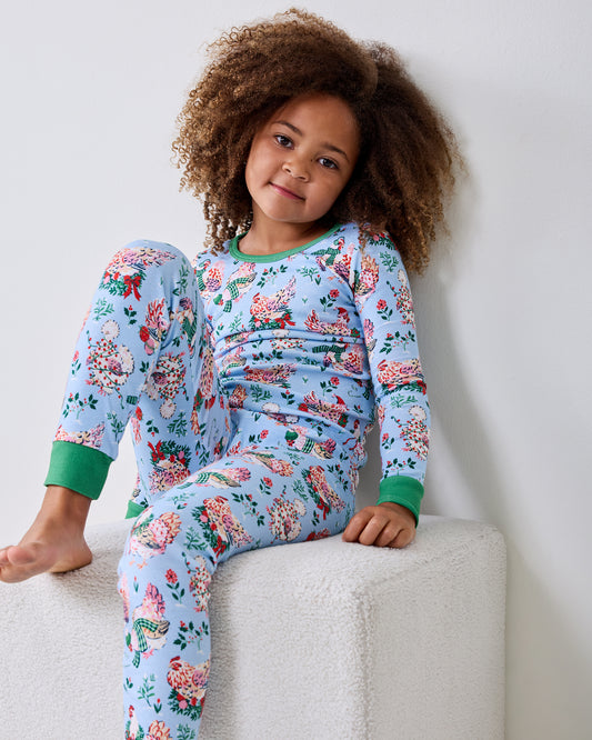 Child wearing a pale blue long-sleeve Holiday Hens pajama set with a floral and hen print, seated on a white ottoman.